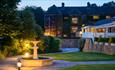 Outdoor area with some lawn and patio. A fountain stands  in the middle of the patio area with small floor lights either side.