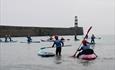 Group of people paddleboarding and kayaking near a lighthouse on a calm sea, wearing helmets and safety vests.