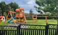 Play area with large wooden equipment including swings, slide, climbing wall and rope walk. Situated in a large grassed area with fencing all around.