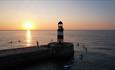 Paddleboarders gather near a lighthouse at sunset, with golden light reflecting on the calm sea.