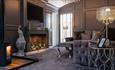 A grey coloured living area with panelled walls, grand fireplace, TV on the wall and log fire burning.