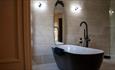 Floor to ceiling tiled bathroom in natural colours with freestanding black bath and a long oval mirror in front.