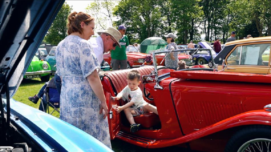 A little boy sits in the back of a classic car with the door open. Families in the background browse the display of cars.