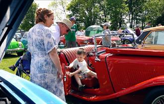 A little boy sits in the back of a classic car with the door open. Families in the background browse the display of cars.