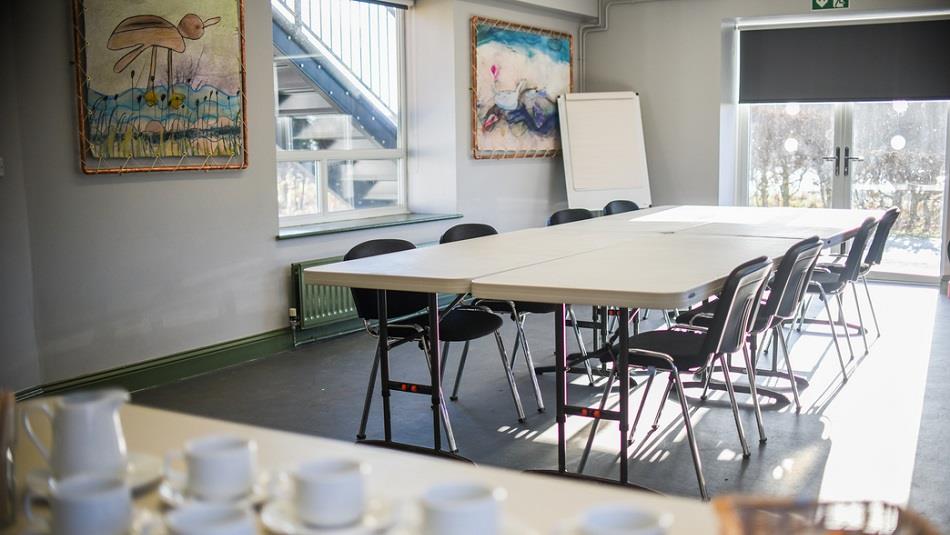 Meeting room with large table surrounded by 8 chairs, flipchart, two paintings on wall, tea cups with milk jug on separate table.  One window and glas