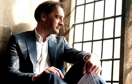 Image of Alistair McGowan sitting by a window holding sheet music - The Piano Show (Funny Way To Be Comedy)