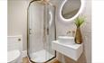 Bathroom with beige colour, textured tiles. A gold shower unit with gold shower head too.
White toilet and sink with gold fixtures and a circular mirr