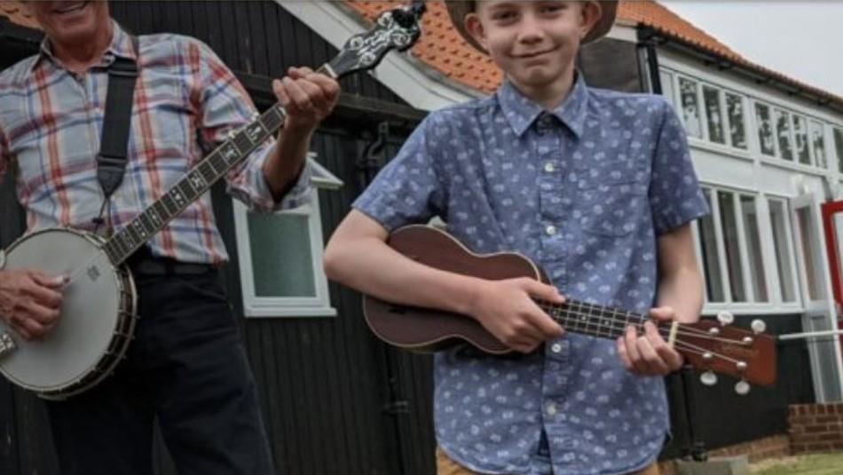 Young boy in blue and white shirt playing a brown ukulele, next to man in checked shirt playing a banjo