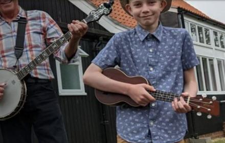 Young boy in blue and white shirt playing a brown ukulele, next to man in checked shirt playing a banjo