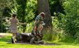 Two children playing in Ushaw’s gardens on a sunny day; one child climbs a carved animal sculpture while the other runs nearby on the grass surrounded
