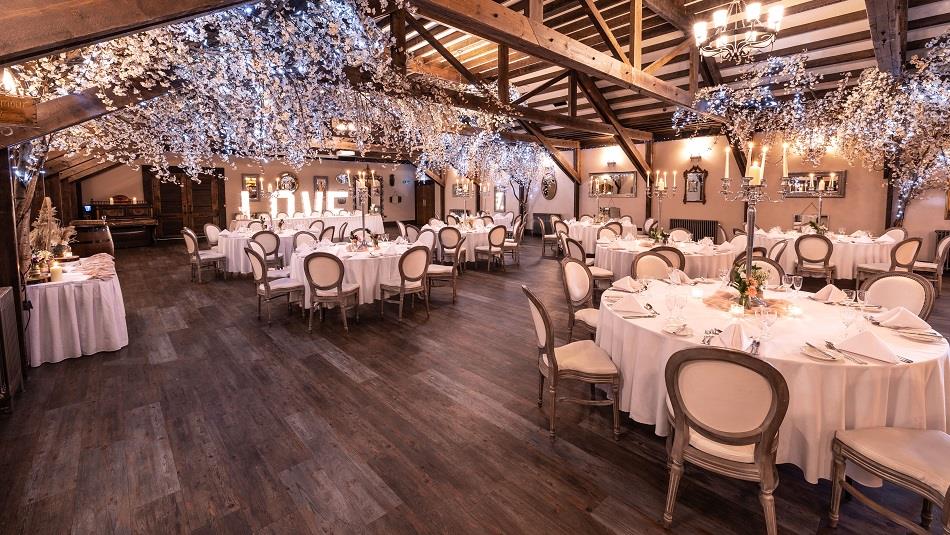 Image of a beautiful dining room decorated for a wedding: softly lit, with several white tables and pretty indoor trees.