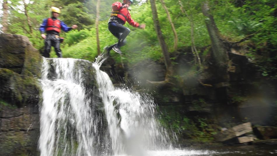 2 children in life vests and safety helmets jumping from some rocks into a river with a small waterfall below them