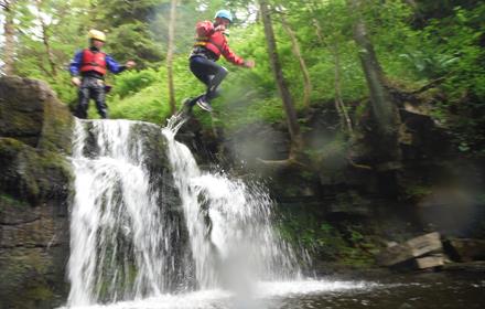 2 children in life vests and safety helmets jumping from some rocks into a river with a small waterfall below them