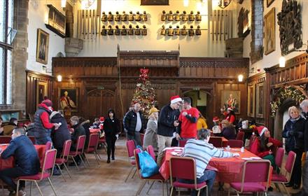 People in Durham castle great hall, tables with red table cloths, chairs, Christmas tree