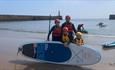 Family of 4 adults and 2 children posing for a photograph on the beach at Seaham marina. Holding a large paddleboard and wearing helmets and lifejacke