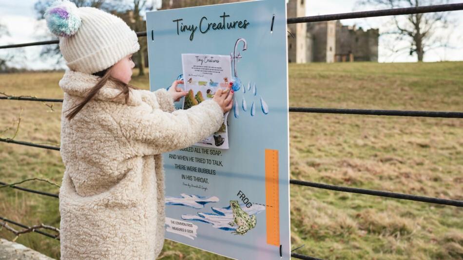 Girl in fluffy coat and woolly hat holding info sheet and looking at a trail poster in Raby Castle's Deer Park