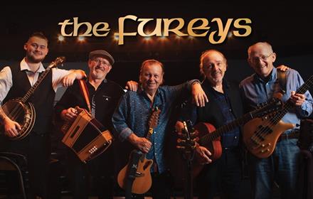 Band members of 'The Fureys' stand together with their instruments, whilst resting their left arm on each other.