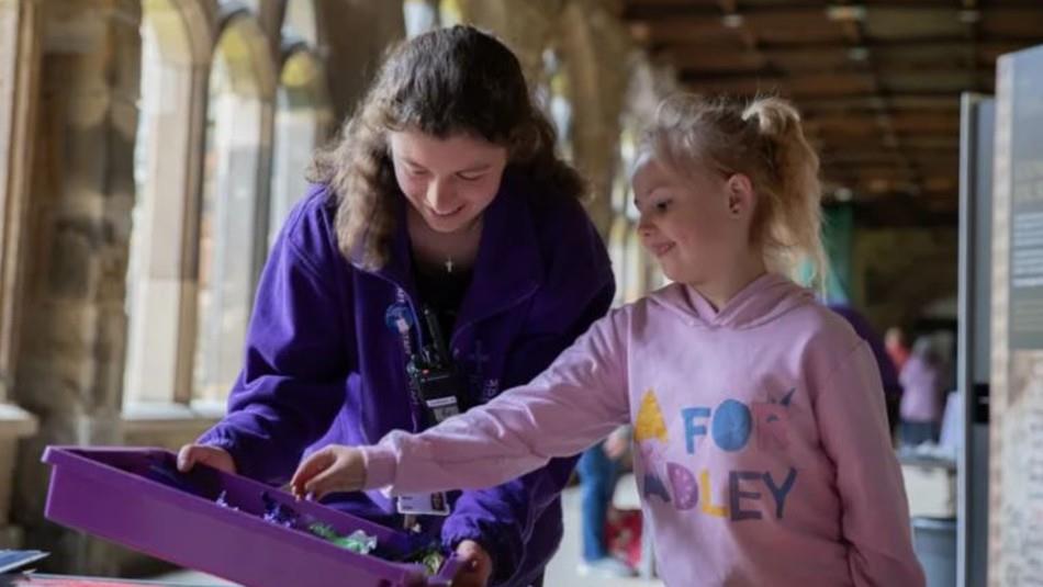 Girl in pink hoodie taking something out of a purple tray held by Durham Cathedral staff member