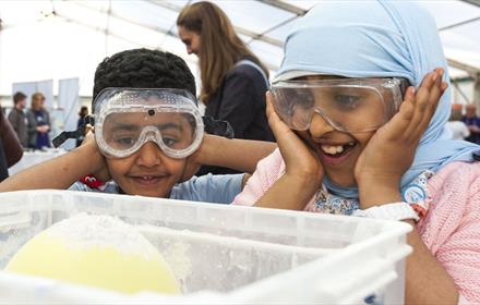 Two smiling children wearing protective goggles, with their hands over their ears excitedly anticipating the climax of an experiment.