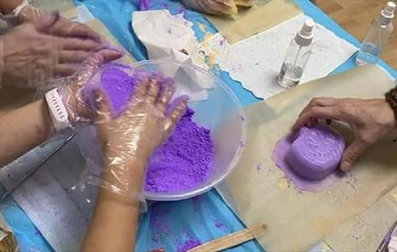 Bath bombs being hand made.  Purple mixture in glass bowl being mixed by hand with a completed bath bomb beside it.