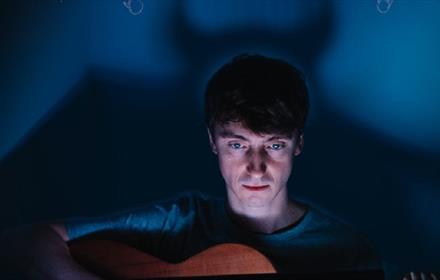 Image of Jonluke McKie playing the guitar on a darkened set with a silhouette of the devil behind him.