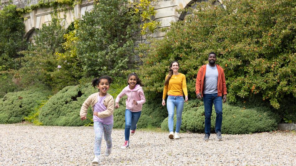 A family, 2 adults and 2 children, running in the grounds of The Bowes Museum, smiling.