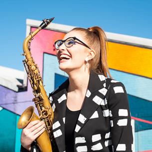 A woman holding a saxophone, in front of a geometric background. 