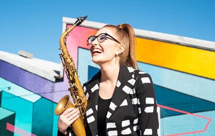 A woman holding a saxophone, in front of a geometric background.