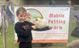 Child holding a white chicken in front of a sign with wording Mobile Petting Farm.