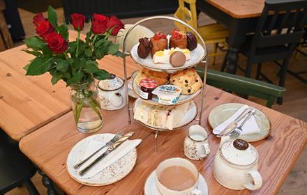 Afternoon tea stand with cakes, sandwiches and scones, red roses, tea cups, teapot and plates.