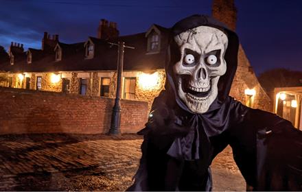person dressed in Halloween costume featuring large skull stood in front of miners cottages at Beamish Museum at night time.