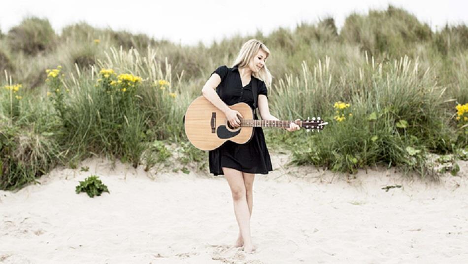 Hattie Murdoch playing the guitar barefoot on the beach, grassy dunes with yellow flowers make up the background.