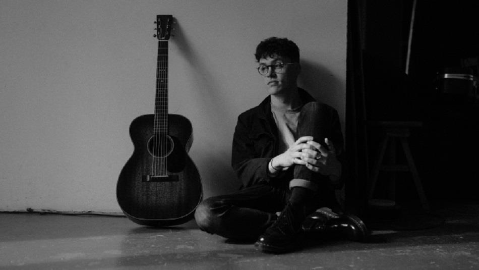Black and white photo of John Gatenby sitting against a wall with his knees drawn up, guitar beside him.