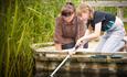 Mum and daughter on wood platform next to a pond. Daughter is holding a net in the water to pond dip for creatures.