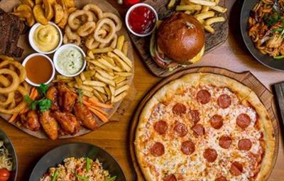 Table with several dishes of food, such as pizza, burger, pasta, Asian dishes and platters.