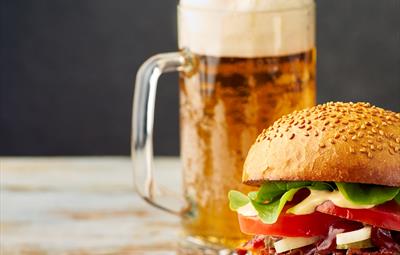 A pint of beer next to a burger filled with lettuce, tomatoes and cheese.
