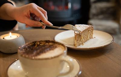 Person holding a fork cutting into a slice of cake on a white plate. White cup of coffee