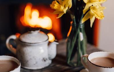 Two white cups of tea, white with black dots teapot. Glass vase with daffodils. Fire.