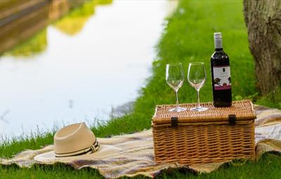 Picnic basket with wine glasses and wine bottle on top. Hat on blanket. Next to body of water.