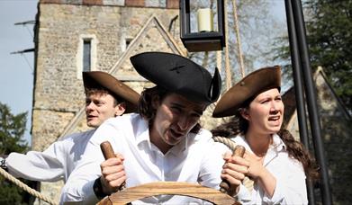 Three actors dressed as sailors stand behind a ships wheel