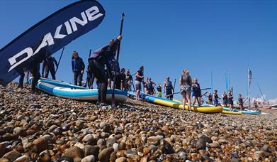 People preparing large paddleboards on a pebbled beach under a clear blue sky.