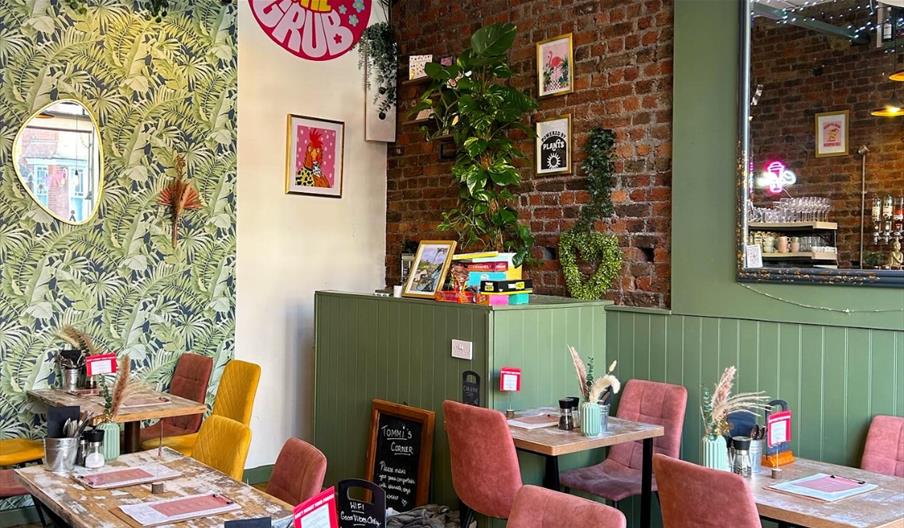 Interior of The Good Grub restaurant with green leaf patterned wallpaper, exposed bricks and wooden tables with yellow and pink chairs.