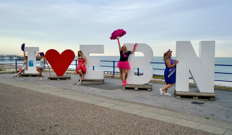 Performers from the Dance Through the Decades show in outfits from various eras posing by the I Heart Eastbourne sign.