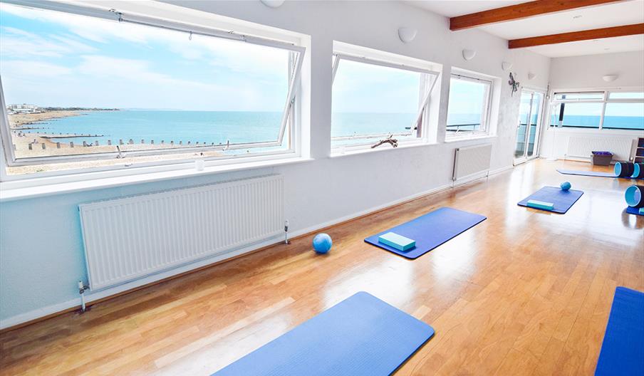 Open plan yoga studio with white walls and wooden flooring with windows offering views of the sea and blue yoga mats spaced out on the floor