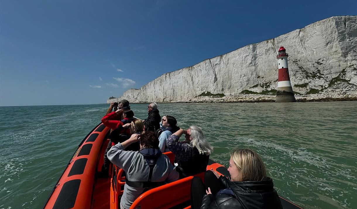 Photo take from back of an orange rib boat, looking over the passengers towards white chalk cliffs and red and white striped beachy head lighthouse