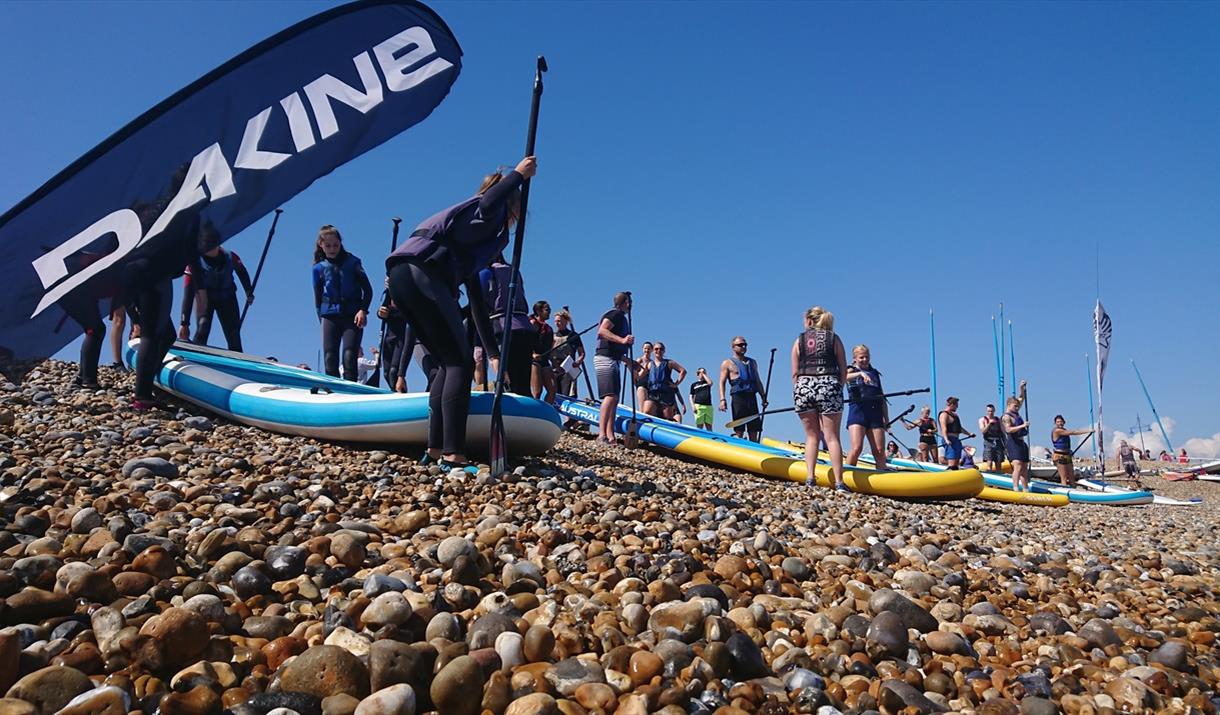 People preparing large paddleboards on a pebbled beach under a clear blue sky.