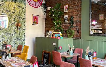 Interior of The Good Grub restaurant with green leaf patterned wallpaper, exposed bricks and wooden tables with yellow and pink chairs.