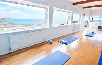 Open plan yoga studio with white walls and wooden flooring with windows offering views of the sea and blue yoga mats spaced out on the floor