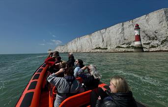Photo take from back of an orange rib boat, looking over the passengers towards white chalk cliffs and red and white striped beachy head lighthouse