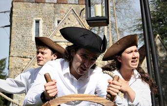 Three actors dressed as sailors stand behind a ships wheel